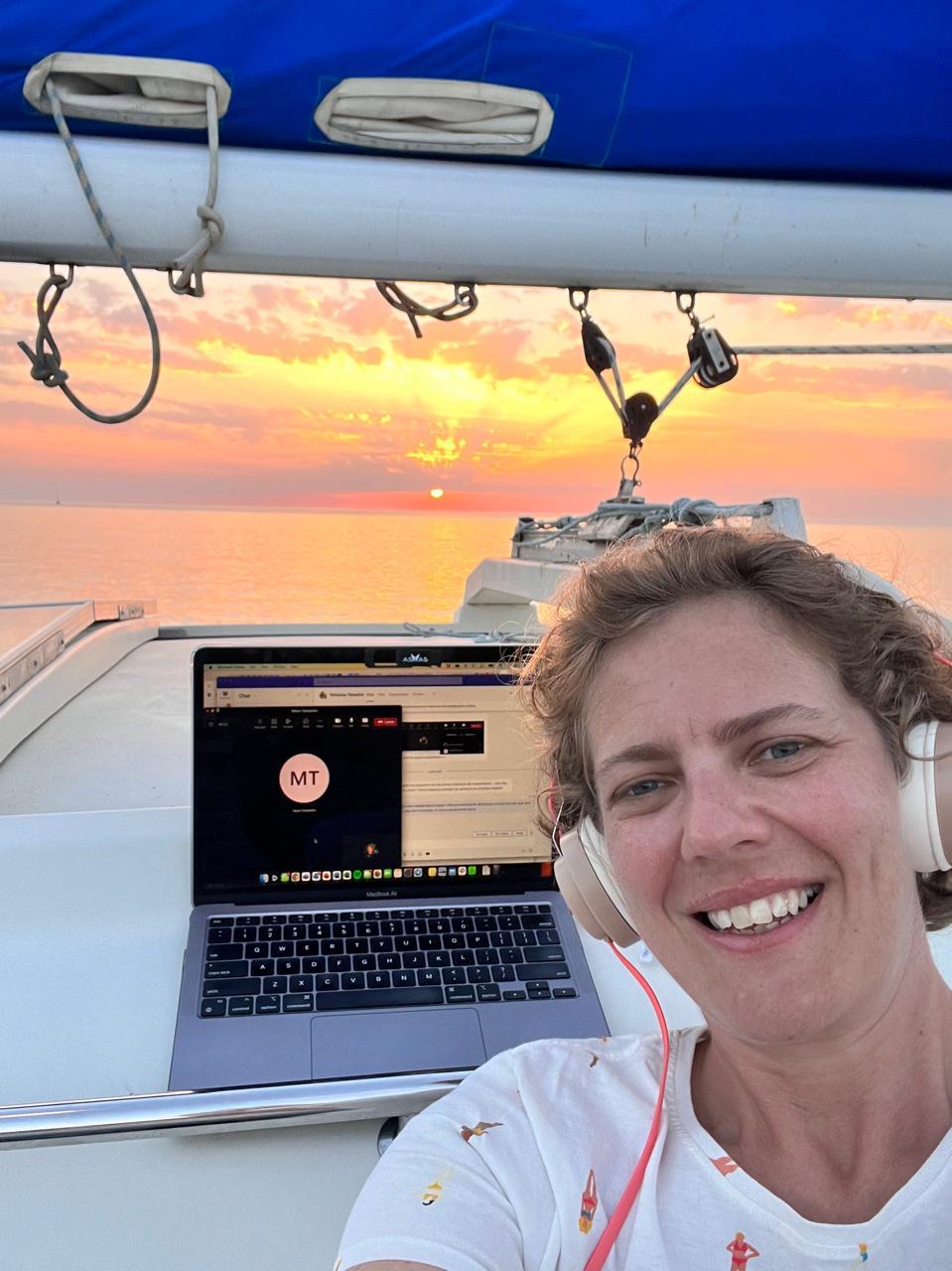anywhere office Mulher branca de fones de ouvido e cabelo curto, sorrindo para a câmera. Atrás dela, computador com tela de reunião e por do sol ao longe, com vista do mar